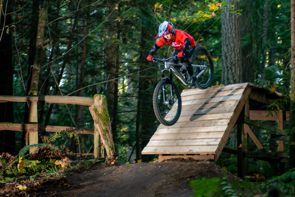 Person on a mountain bike performing a jump over a wooden ramp in a forest setting